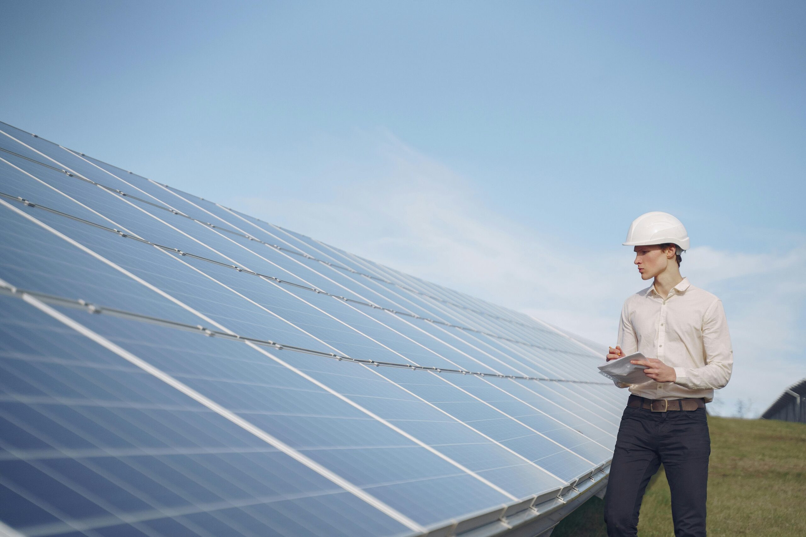 Young serious male engineer in formal outfit and helmet reporting while inspecting solar panel field